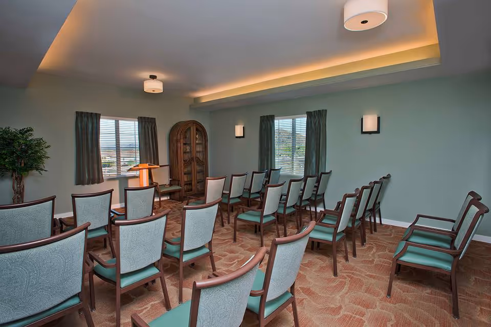 A small meeting or presentation room with rows of wooden chairs with green cushions arranged facing a wooden podium. The room has light green walls, two windows with dark curtains, a wooden cabinet in the corner, a potted plant, and ceiling lights with soft yellow accent lighting.