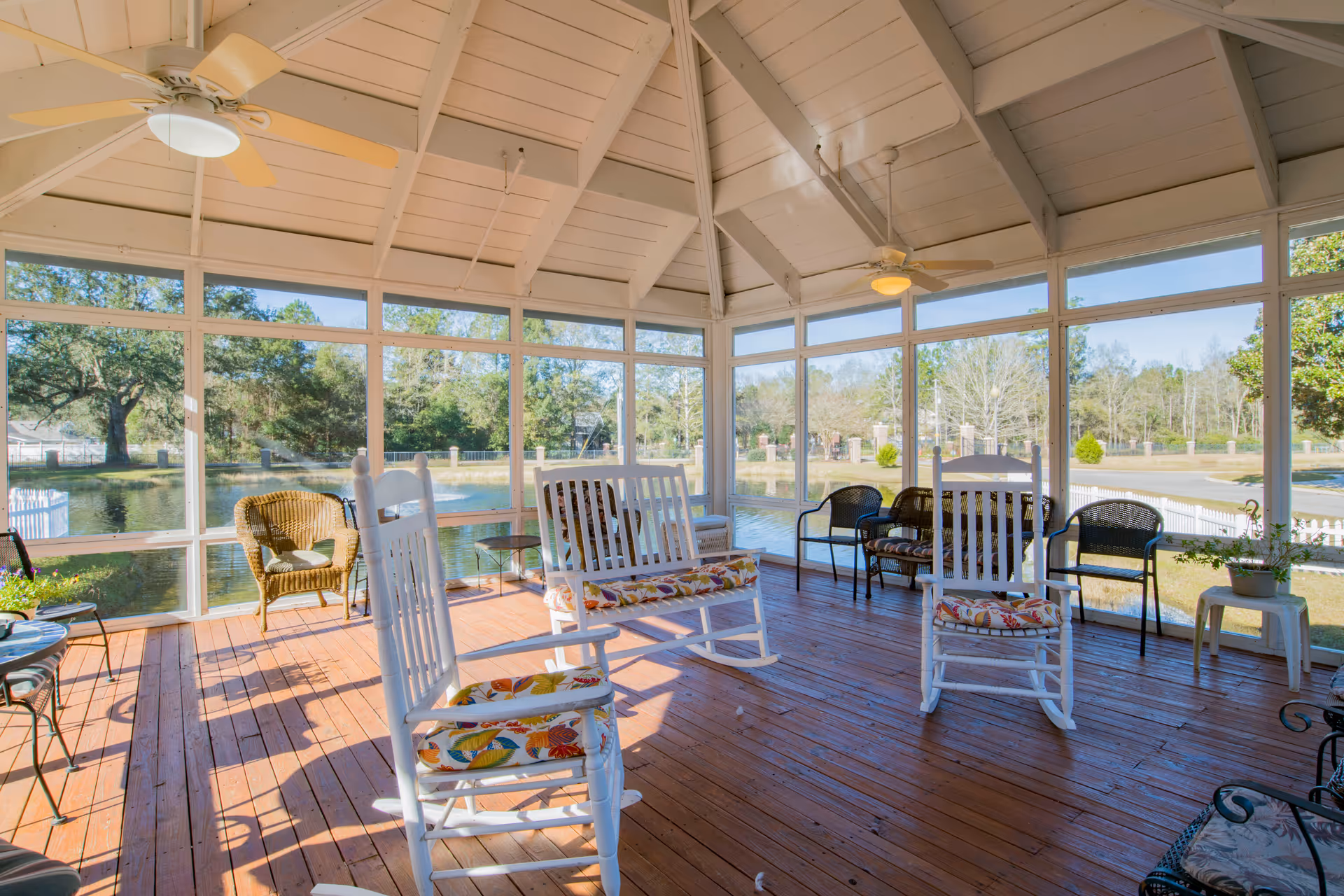 A bright, screened-in porch with wooden flooring and a vaulted ceiling with two ceiling fans. The porch is furnished with white rocking chairs with floral cushions, wicker chairs, and metal chairs. Large windows provide a view of a pond and trees outside.