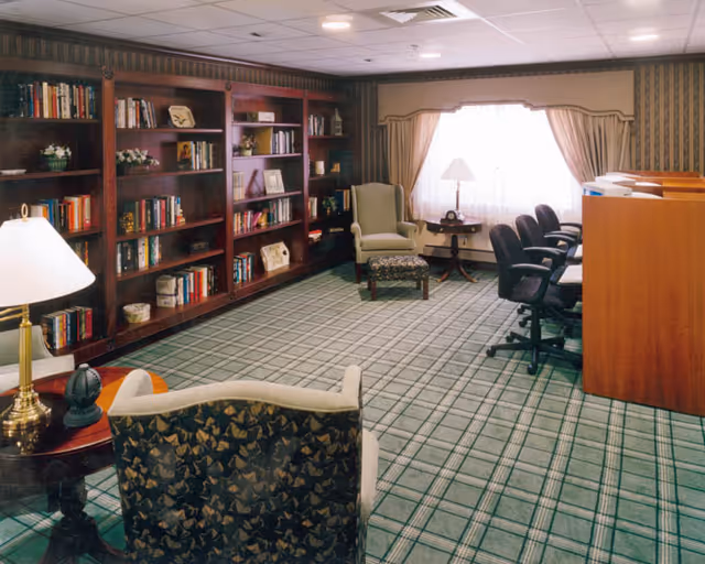 A cozy room with a large wooden bookshelf filled with books and decorative items along one wall. The room has patterned carpet flooring and wallpapered walls. There are several chairs including an armchair with a matching ottoman, a small round table with a lamp, and a row of office chairs facing wooden cubicles. A large window with curtains allows natural light into the room.