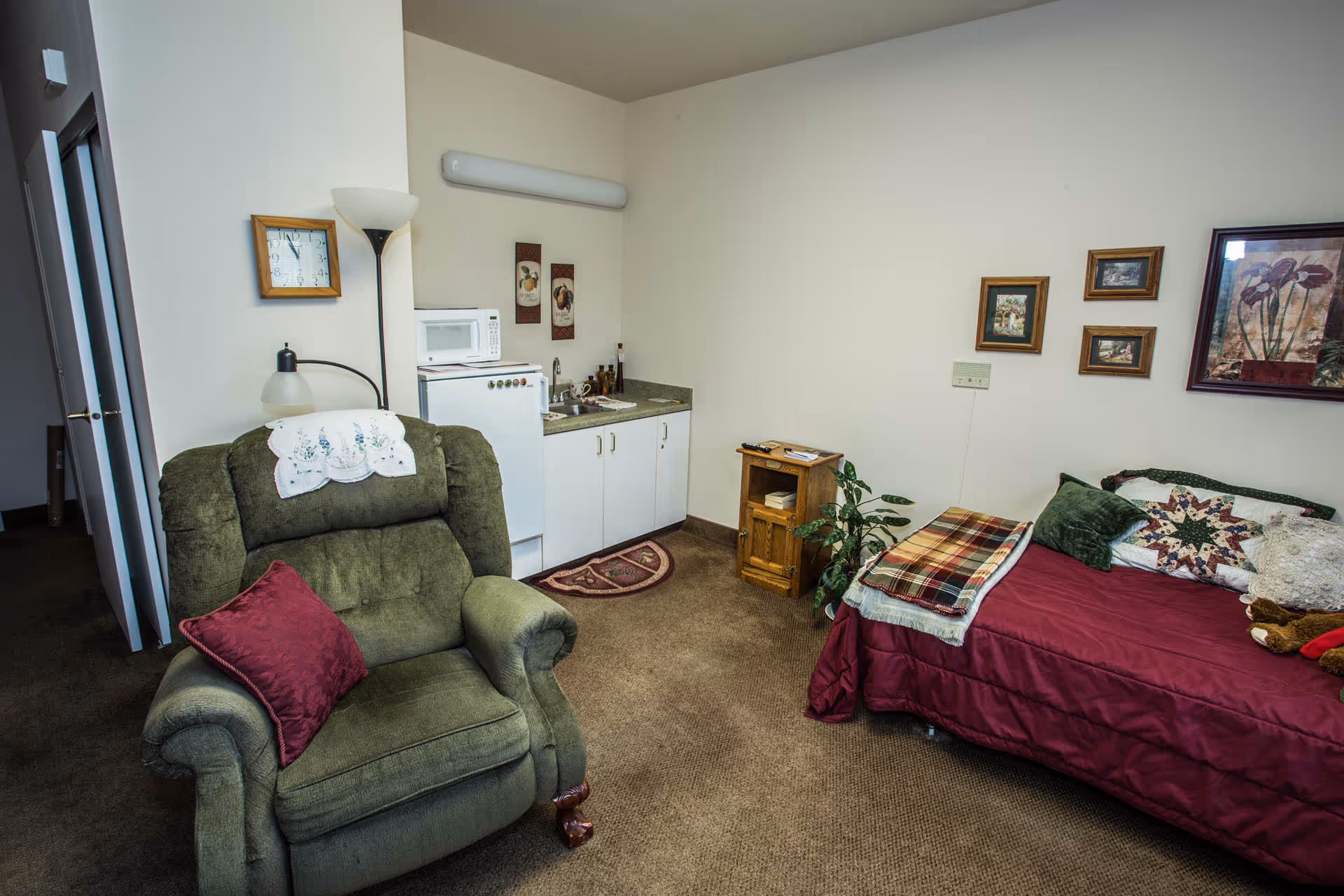 A cozy assisted living room featuring a green upholstered armchair with a red pillow and a white doily on the backrest, a small kitchenette with a mini fridge, microwave, and sink, a wooden side table with a plant beside it, and a bed with a red comforter, multiple pillows, and a plaid blanket. The walls are decorated with framed pictures and artwork.