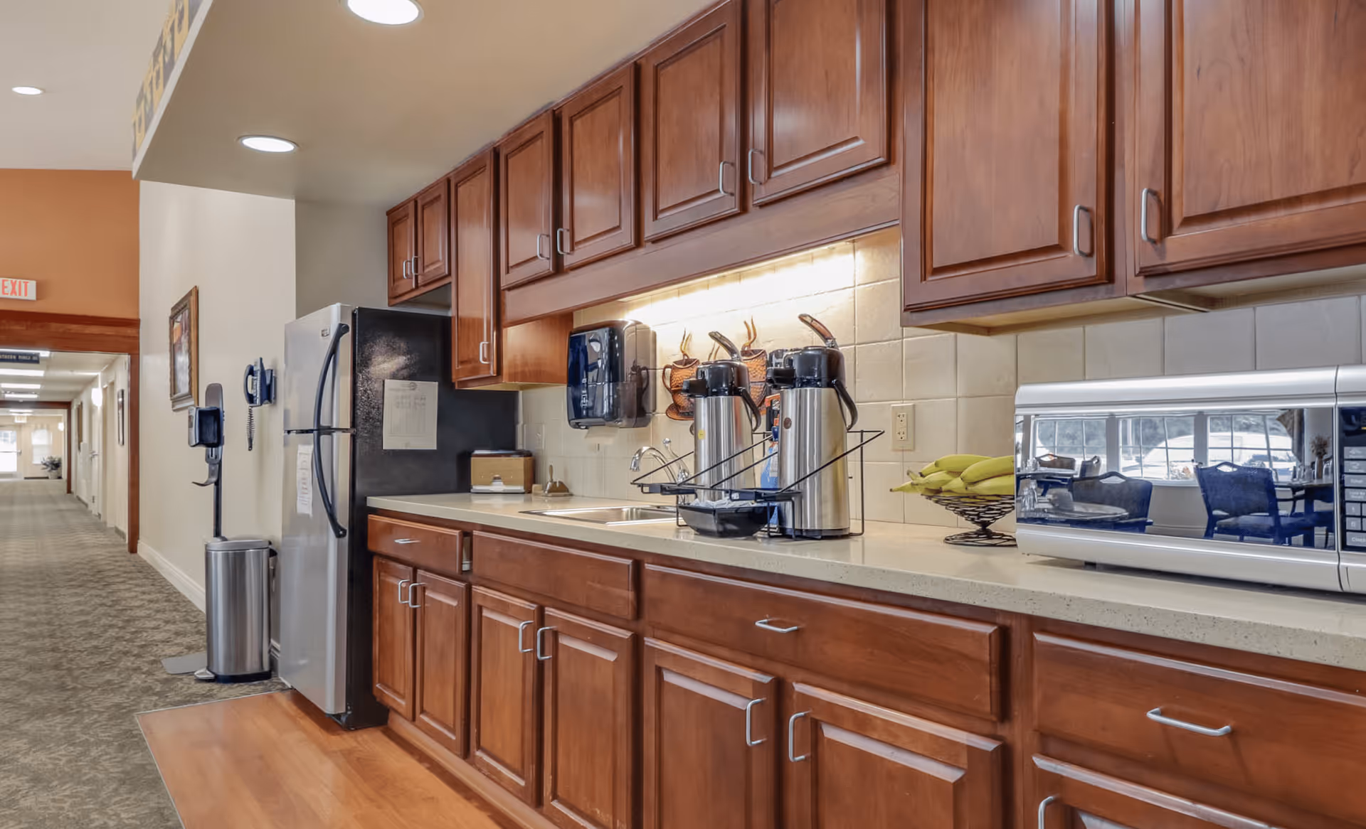 A kitchen area in Arbor Grove Assisted Living & Memory Care featuring wooden cabinets, a countertop with a microwave, coffee dispensers, a fruit basket with bananas, and a stainless steel refrigerator. The kitchen is adjacent to a hallway with carpeted flooring and an exit sign visible in the distance.
