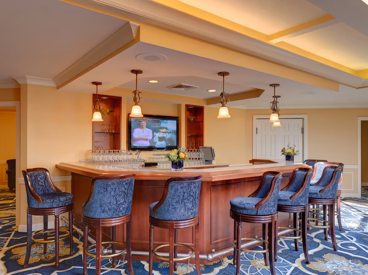 Elegant indoor bar area with a wooden counter, upholstered bar stools, pendant lights and a wall-mounted TV.