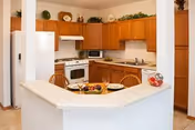 A clean and well-lit kitchen with wooden cabinets, a white refrigerator, stove, microwave, and a countertop with a bowl of fruit and plates. The kitchen is open to a small dining area with a wooden chair visible.