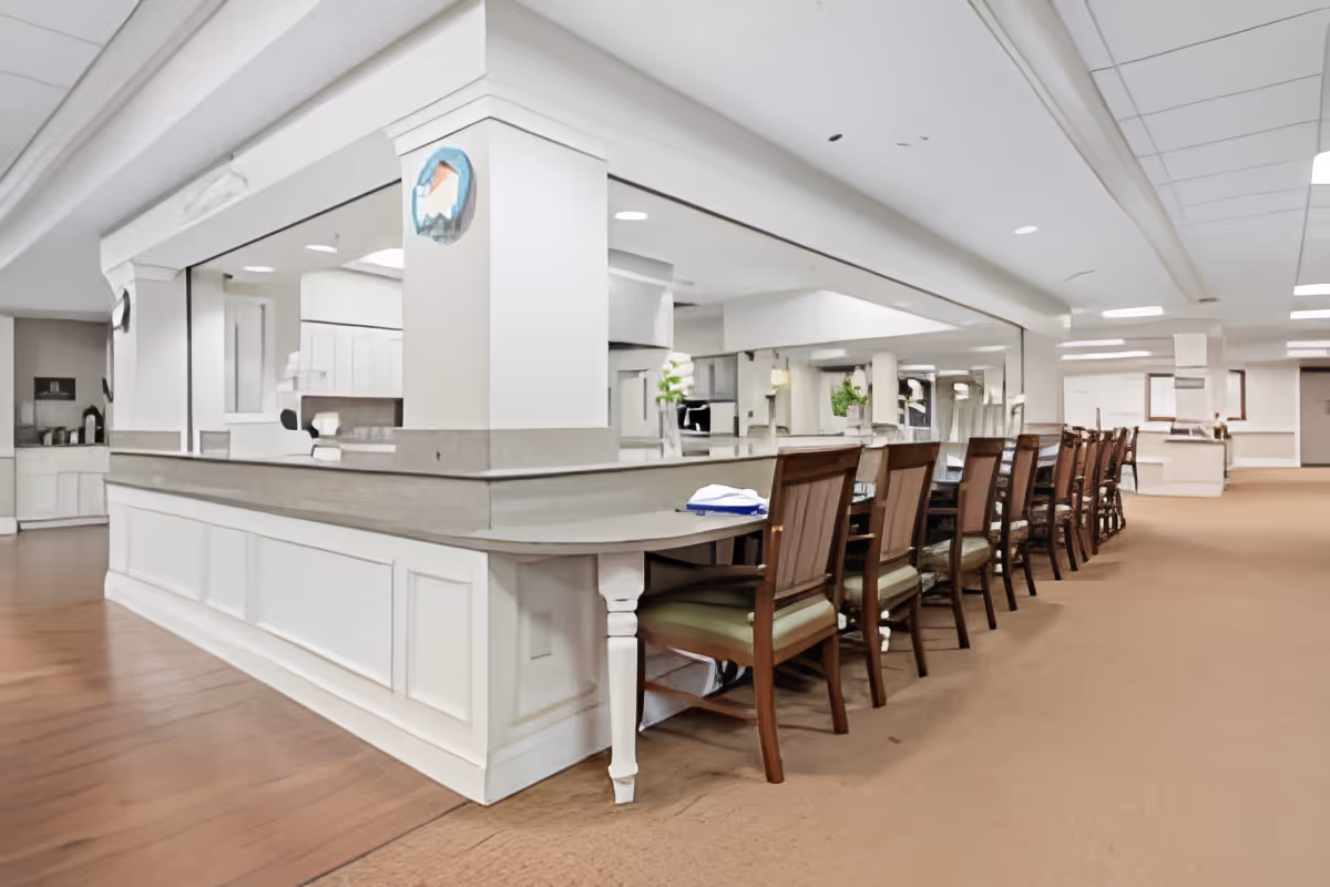 Bright open dining area with a long counter and a row of wooden chairs in a senior living facility.