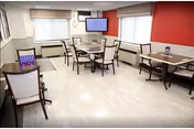 Well-lit dining/activity room with several tables and chairs, light wood flooring, a red accent wall, and a wall-mounted TV.