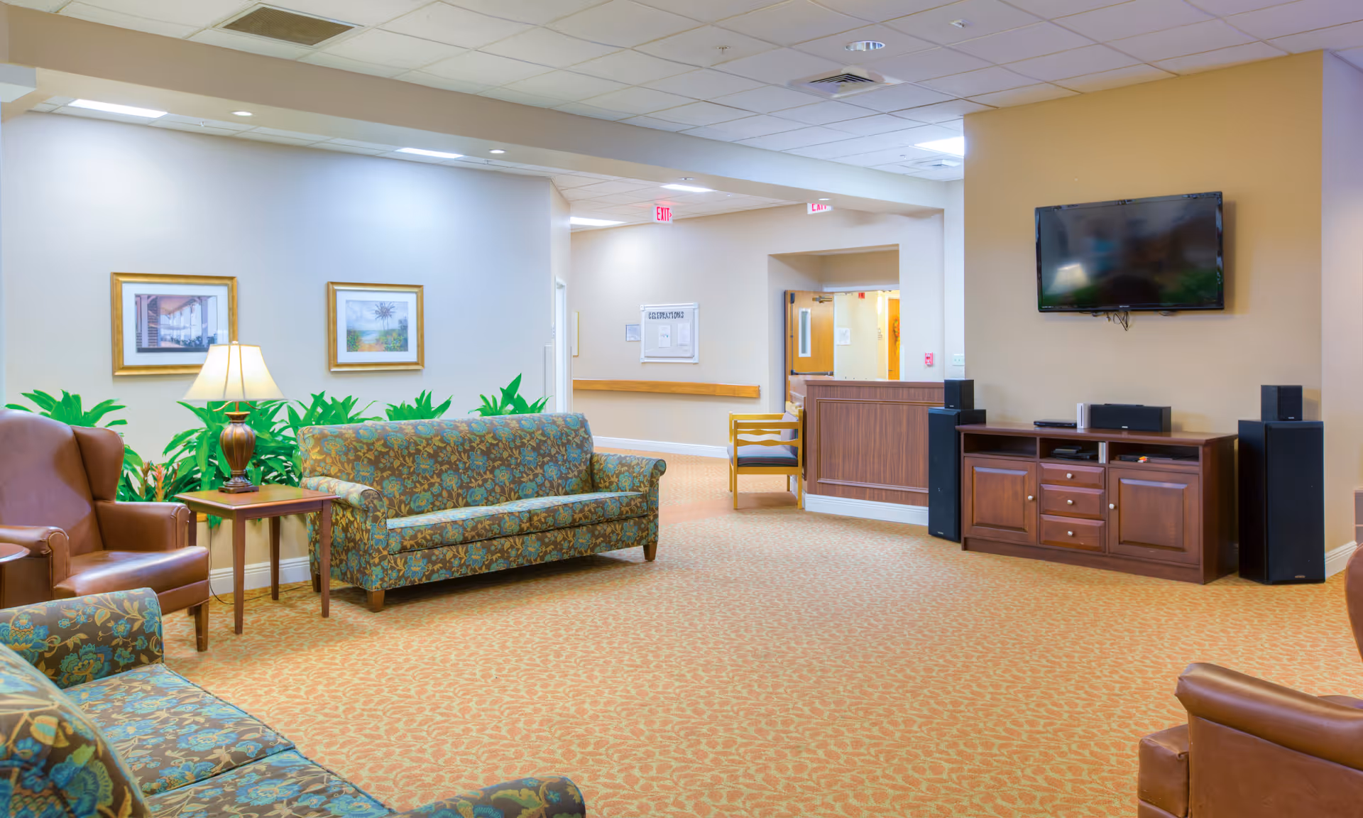 A well-lit senior living facility common area with patterned sofas and leather armchairs arranged around a wooden side table with a lamp. The room has beige walls with framed artwork, a carpeted floor, a wall-mounted flat-screen TV above a wooden cabinet with speakers, and a hallway leading to other rooms.