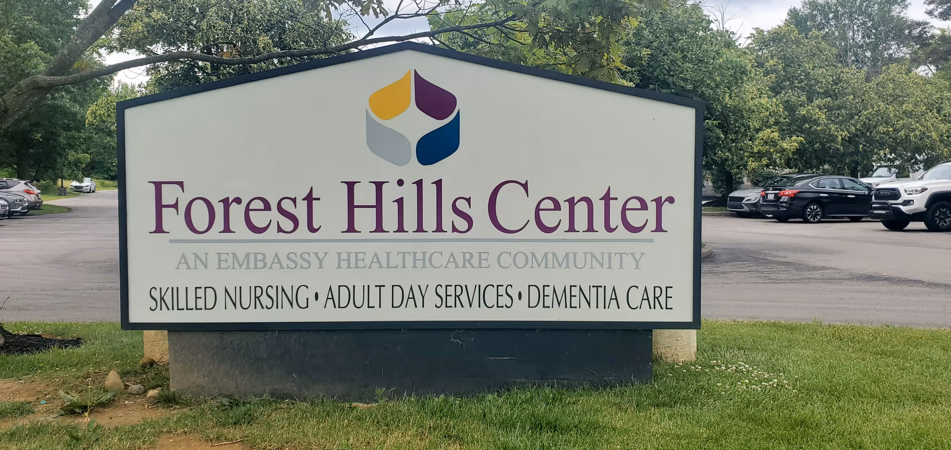 Outdoor sign for Forest Hills Center, an Embassy Healthcare Community, offering skilled nursing, adult day services, and dementia care, with parked cars and trees in the background.