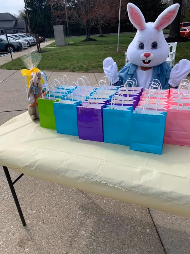 A person dressed in a white Easter Bunny costume with a blue jacket is sitting behind a table covered with a yellow tablecloth. On the table, there are multiple colorful gift bags in green, blue, purple, and pink. To the left of the table, there is a stuffed bunny toy wrapped in clear plastic with a yellow ribbon. The setting is outdoors on a paved area with grass and trees in the background.