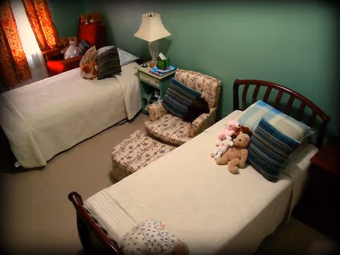 A cozy bedroom with two single beds, each with white bedspreads and striped pillows. One bed has stuffed animals including a teddy bear and a bunny. Between the beds is a small nightstand with a lamp, books, and a box of tissues. There is a cushioned armchair with a matching ottoman next to the nightstand. The room has green walls and a window with patterned curtains.