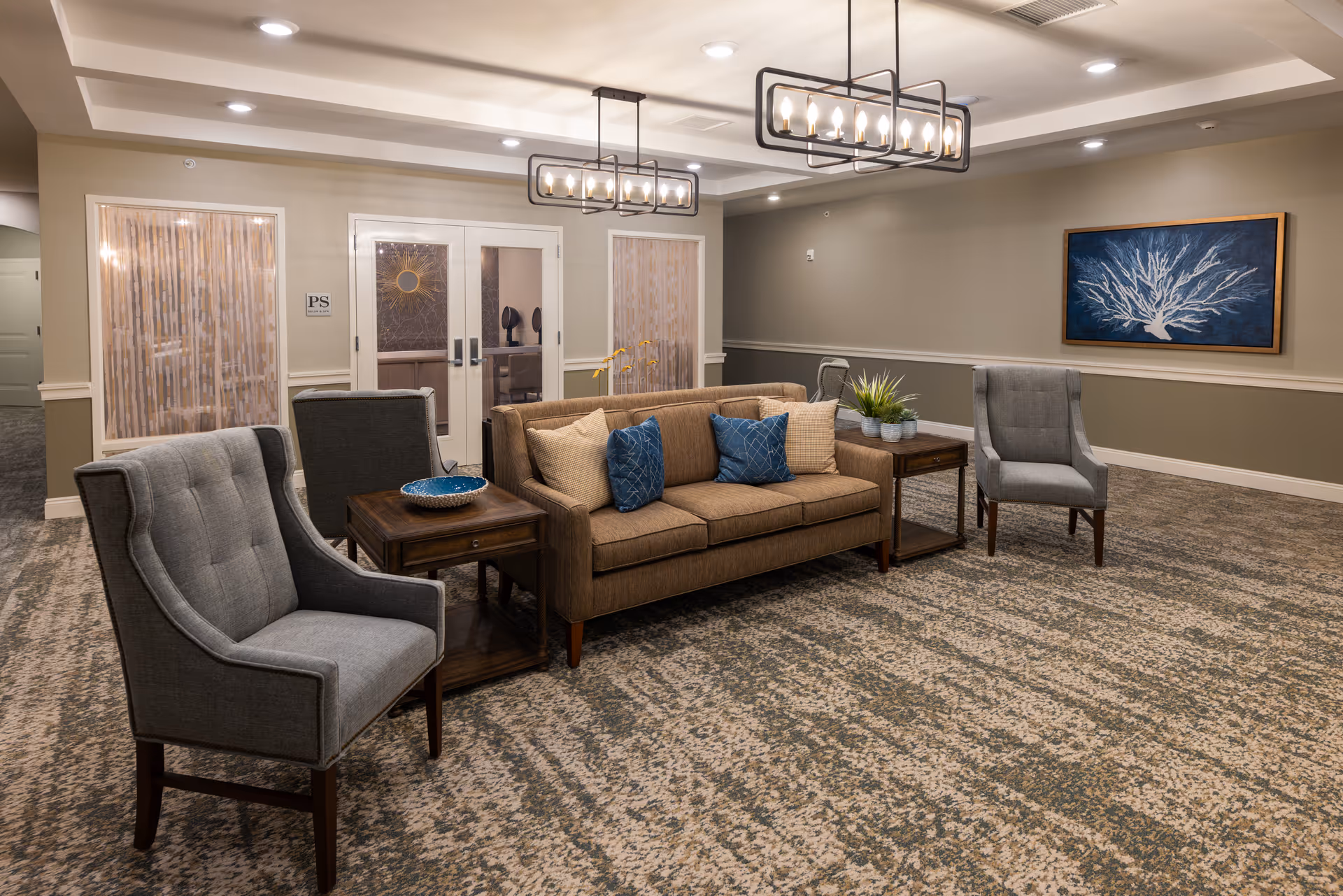 A cozy seating area in a senior living facility featuring a brown sofa with blue and beige pillows, two gray armchairs, and two wooden side tables with decorative items. The room has patterned carpet flooring, beige walls with white trim, modern rectangular pendant lights, and a framed artwork of a white coral on a blue background hanging on the wall.