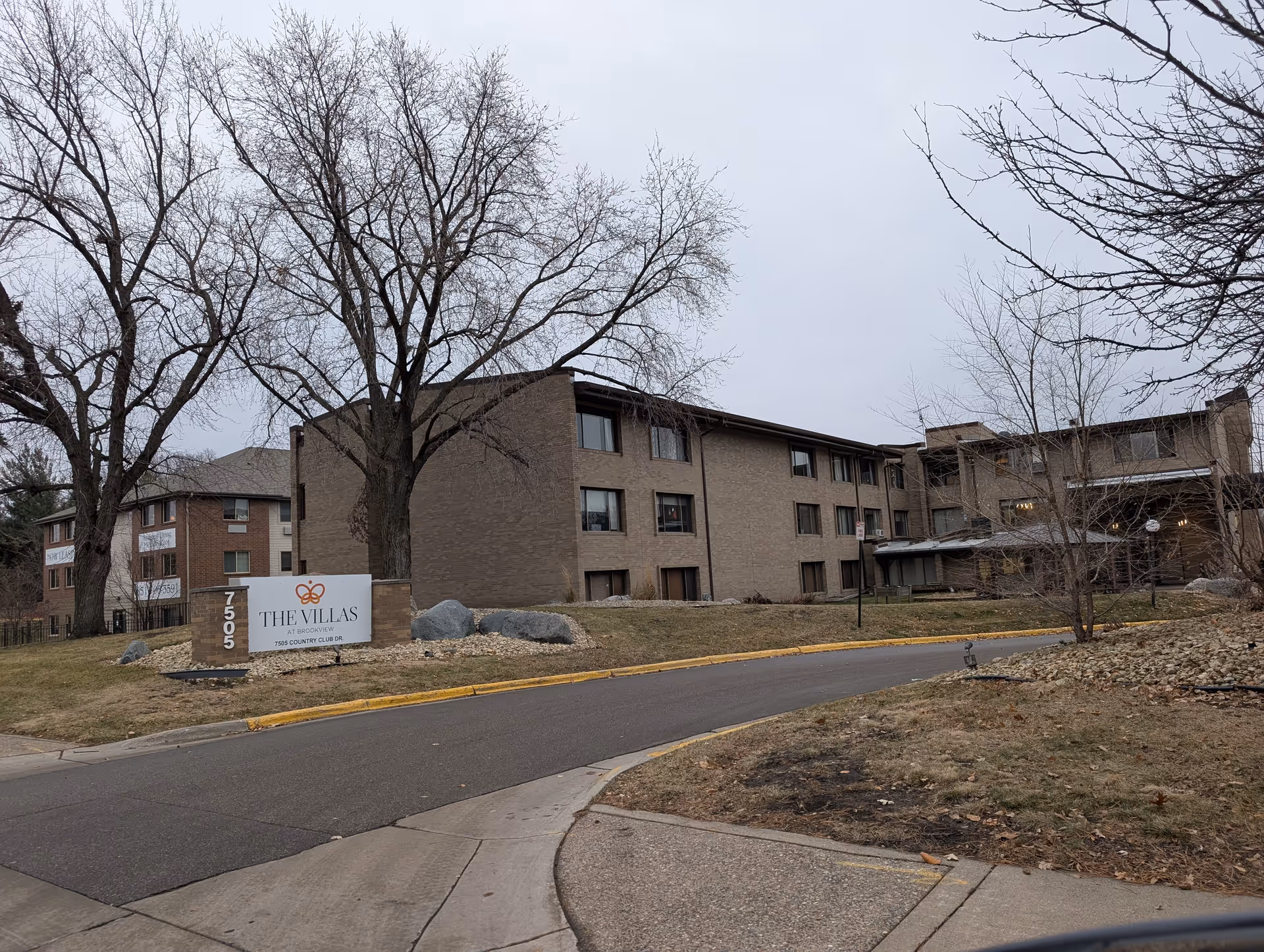 Exterior view of The Villas at Brookview building with a sign displaying the name and address at the front. The building is three stories tall with multiple windows and surrounded by leafless trees and a paved driveway.