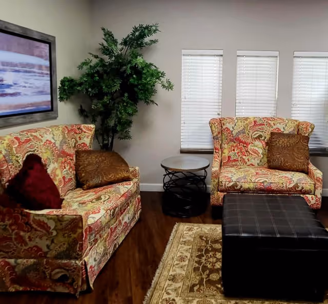 A cozy living room with two patterned armchairs featuring red, yellow, and beige designs, each with decorative pillows. Between the chairs is a small round table with a black metal base and a light-colored top. A large green potted plant is placed in the corner near the wall. The room has three windows with white blinds, a framed abstract painting on the wall, a dark brown ottoman in front of the chairs, and a beige patterned rug on the wooden floor.