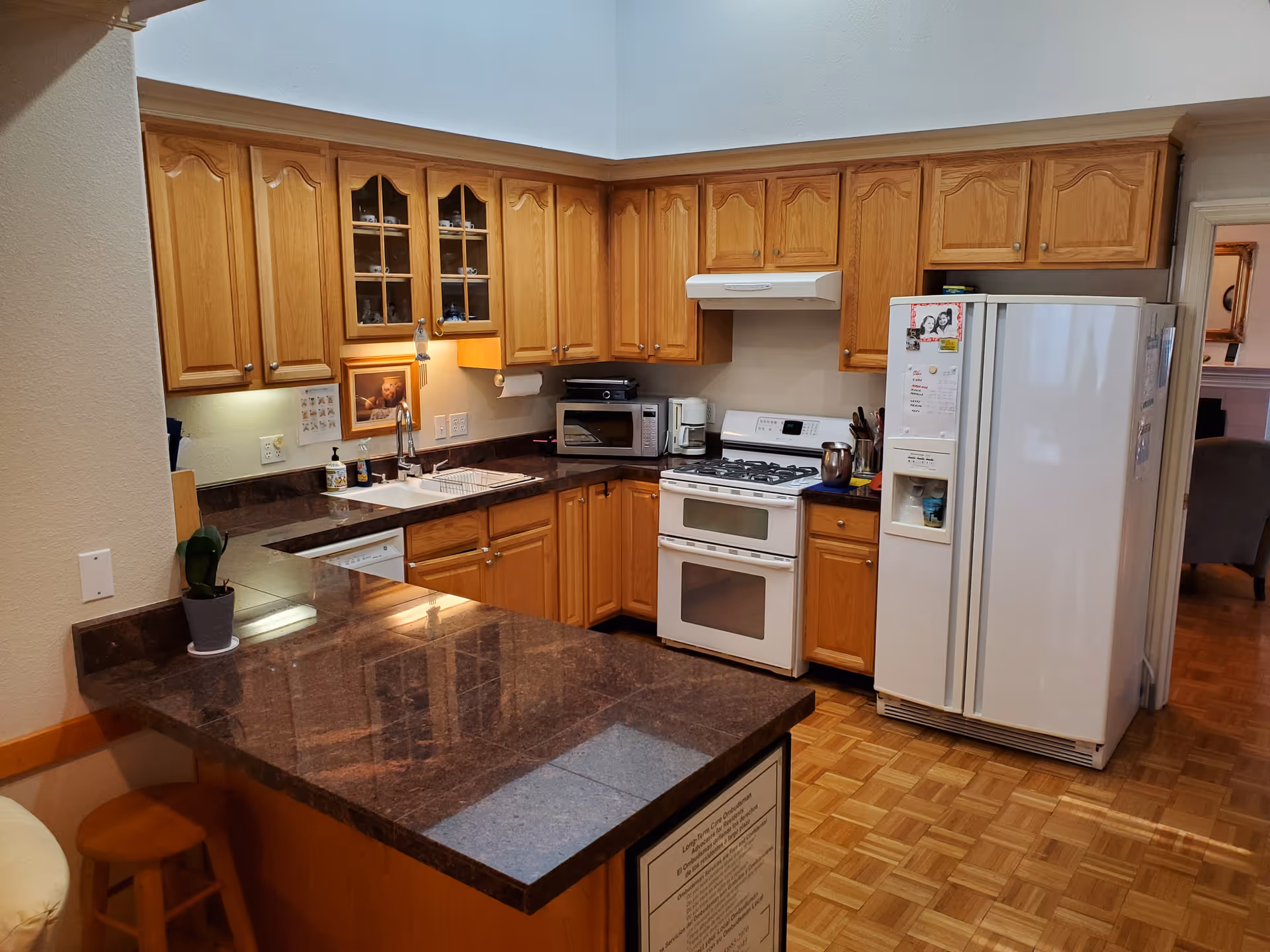 A kitchen with wooden cabinets, a white refrigerator, a white stove with oven, a microwave, a coffee maker, and a sink. The countertops are dark brown granite, and there is a small potted plant on the counter. The floor is parquet wood, and there is a bar stool next to the counter.