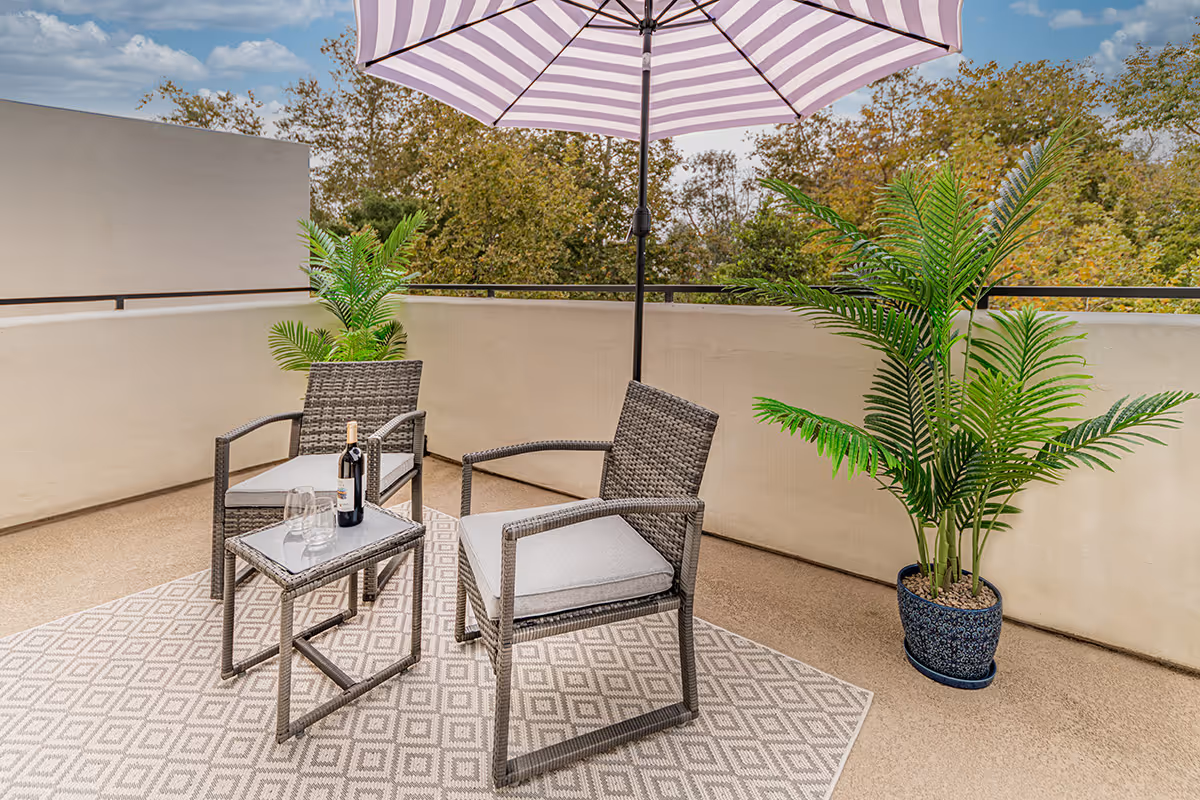A furnished outdoor patio with two wicker chairs, a small table holding a bottle of wine and glasses, a striped umbrella, and potted plants.