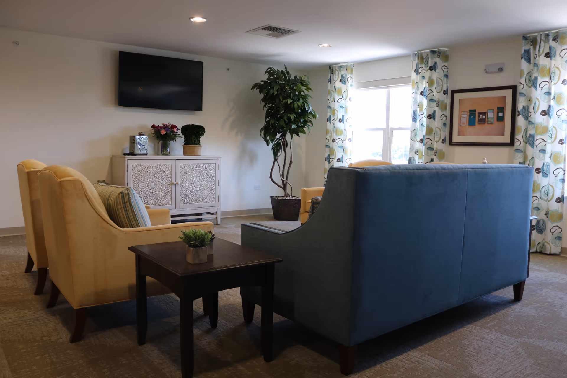 A cozy living room area with a blue sofa and two yellow armchairs arranged around a dark wooden side table with a small plant. A white cabinet with decorative carvings holds a vase of flowers and a small topiary, with a flat-screen TV mounted on the wall above it. A large potted plant stands near a window with patterned curtains, and a framed picture hangs on the wall.