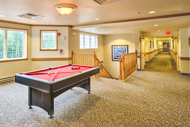 Interior view of a senior living facility common area featuring a red pool table with cues and balls set up. The space has carpeted floors, beige walls with wooden trim, windows letting in natural light, a staircase with wooden railing, and a hallway leading to other rooms.