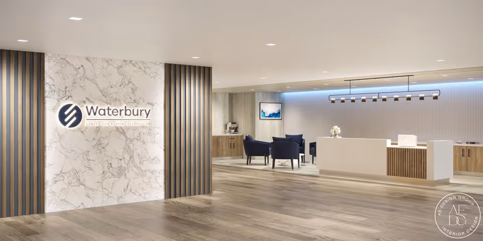 Modern reception area of Waterbury Center for Nursing & Rehabilitation with a marble accent wall featuring the facility's logo, a sleek white reception desk, blue chairs arranged in a seating area, wood flooring, and soft overhead lighting.