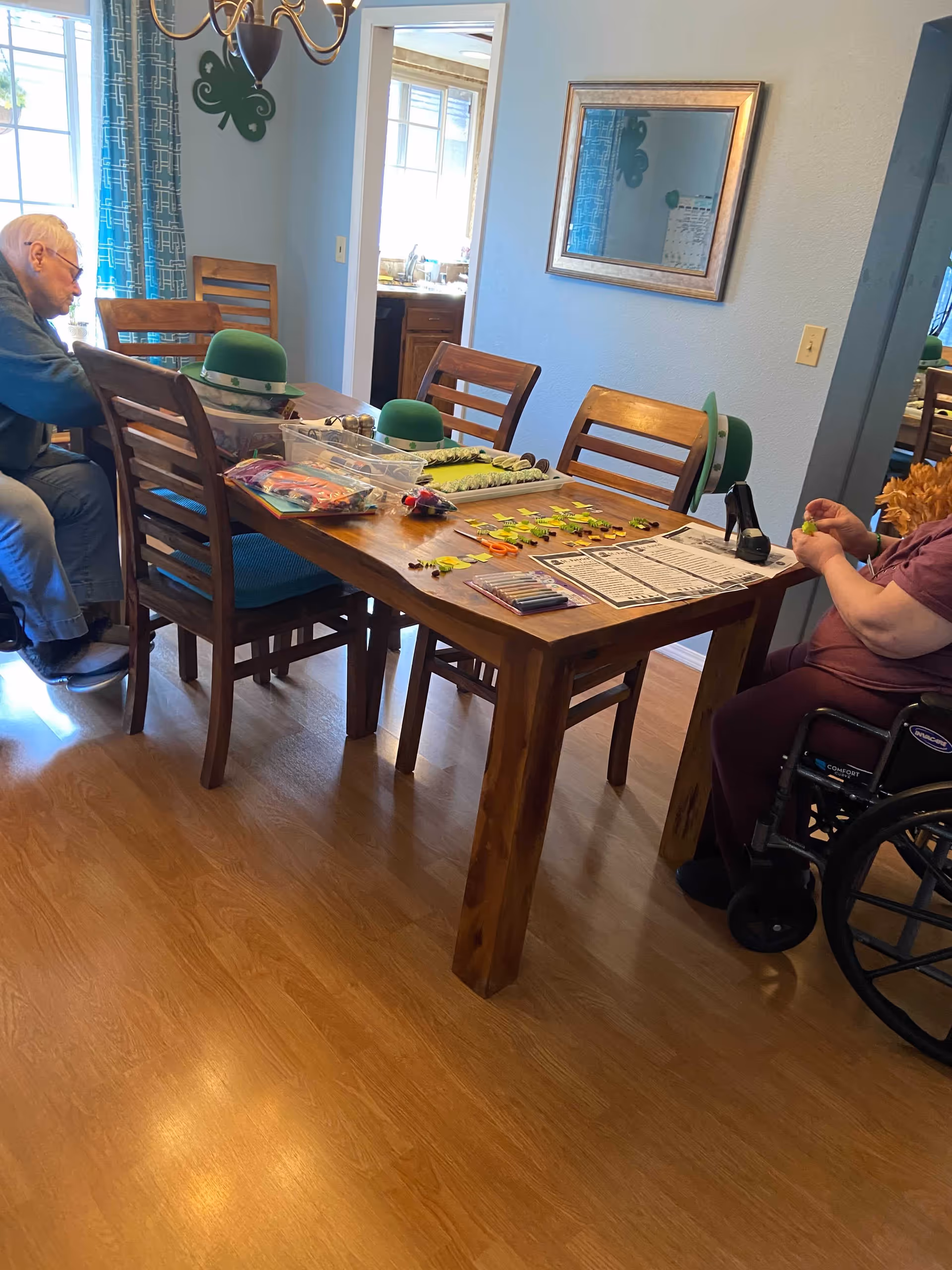 A wooden dining table in a dining room where two people sit and work on arts-and-crafts with green St. Patrick's Day decorations.