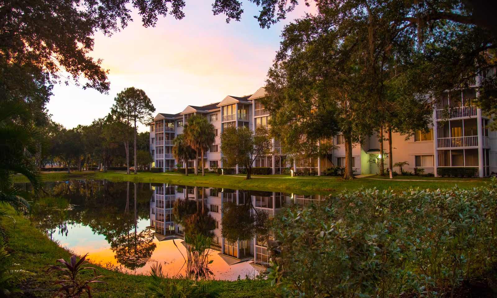 A serene outdoor view of a senior living facility at sunset, featuring a multi-story building with balconies reflected in a calm pond surrounded by lush greenery and trees.