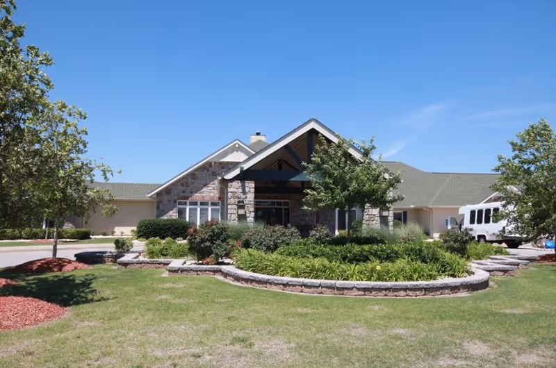 Front exterior view of Homestead Assisted Living & Memory Care of Owasso building with a stone facade, a covered entrance, landscaped garden beds with shrubs and small trees, and a clear blue sky.