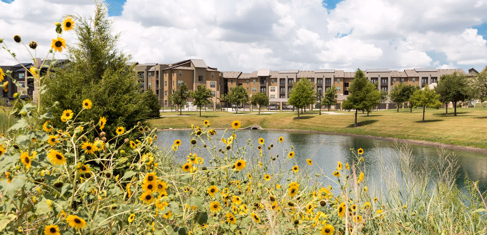 A multi-story residential building seen across a pond with sunflowers and trees in the foreground under a blue sky with clouds.