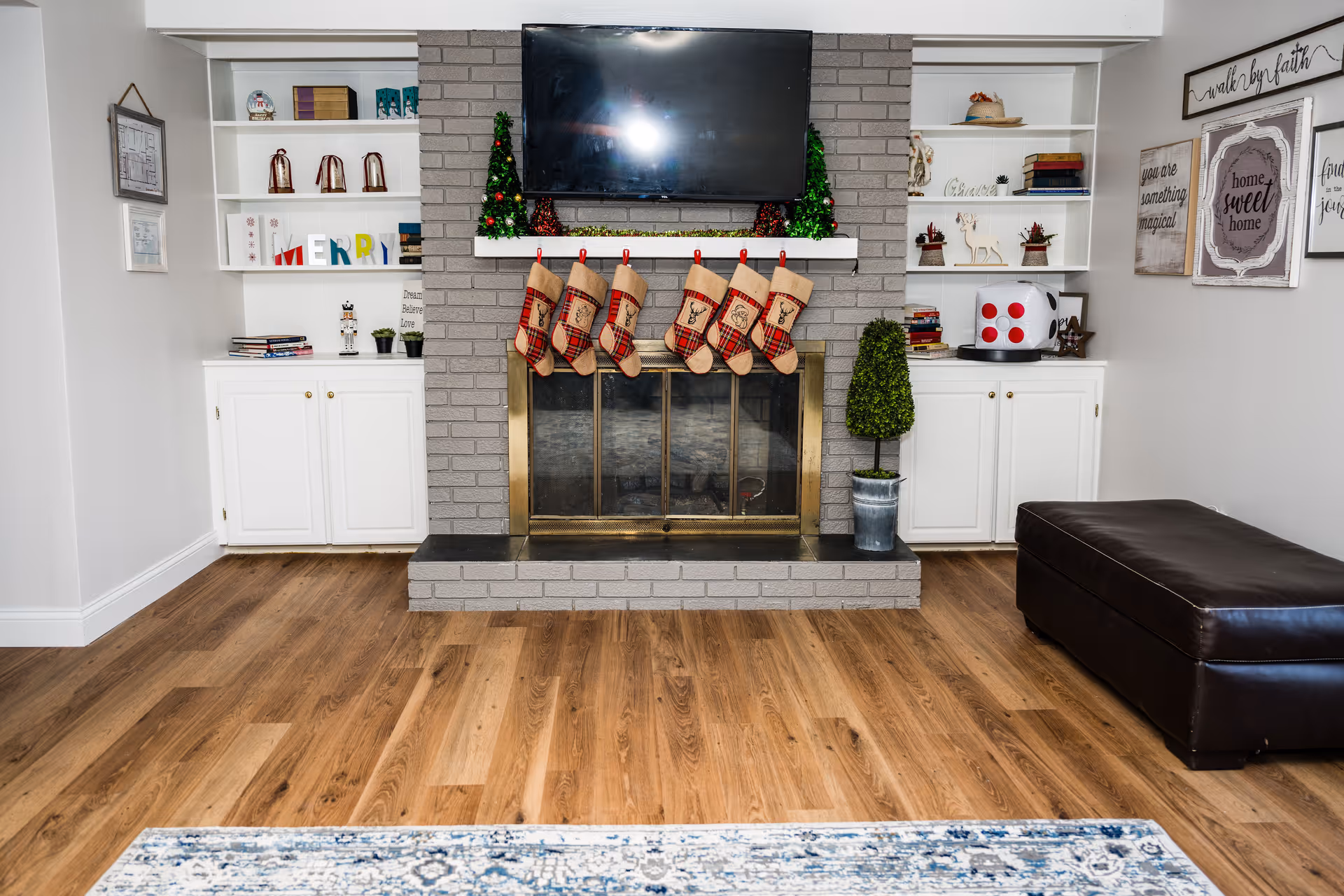 A cozy living room with a gray brick fireplace decorated for Christmas with six red and beige stockings hanging from the mantel. Above the mantel is a flat-screen TV. The mantel also has small Christmas trees and garland. On either side of the fireplace are white built-in shelves and cabinets with various decorative items, books, and holiday decorations. To the right is a dark brown leather ottoman, and a patterned rug is partially visible on the wooden floor. The walls have framed inspirational quotes and signs.