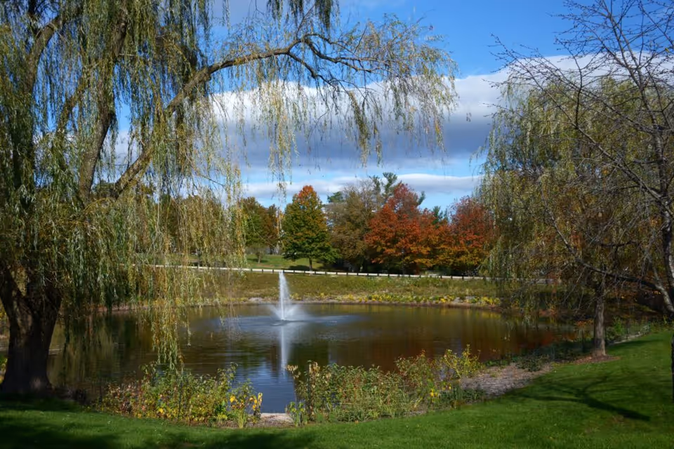 A serene outdoor scene featuring a small pond with a fountain in the center, surrounded by green grass and trees with autumn foliage. The sky is partly cloudy with patches of blue visible.