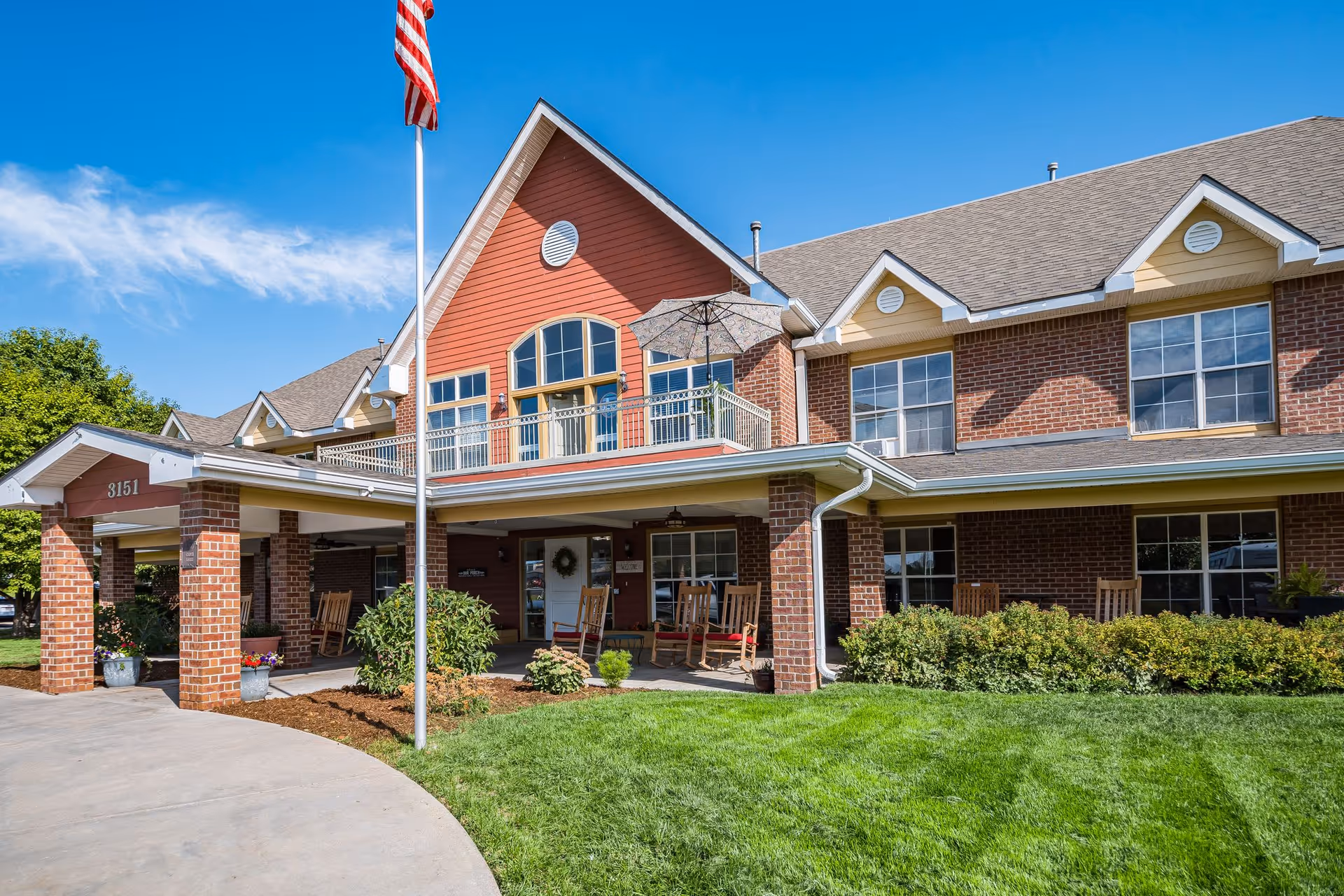 Front exterior view of a two-story assisted living facility with a covered entrance supported by brick columns, rocking chairs on the porch, a balcony with an umbrella, and an American flag on a flagpole in front. The building has red and yellow siding with multiple windows and a well-maintained lawn and shrubs.