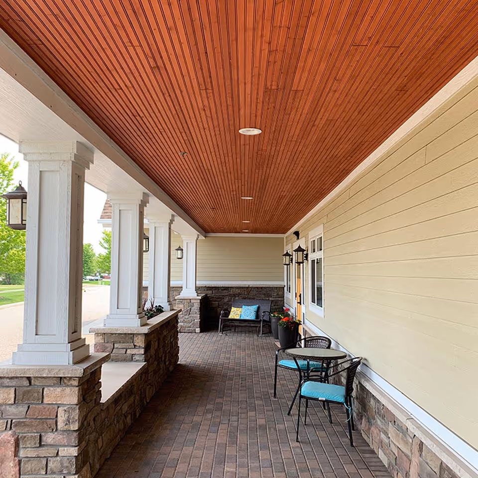 Covered outdoor patio area with brick flooring, stone pillars with white wooden columns, a wooden ceiling, a small round table with two chairs with blue cushions, a bench with colorful pillows, and potted plants along the wall of a building.