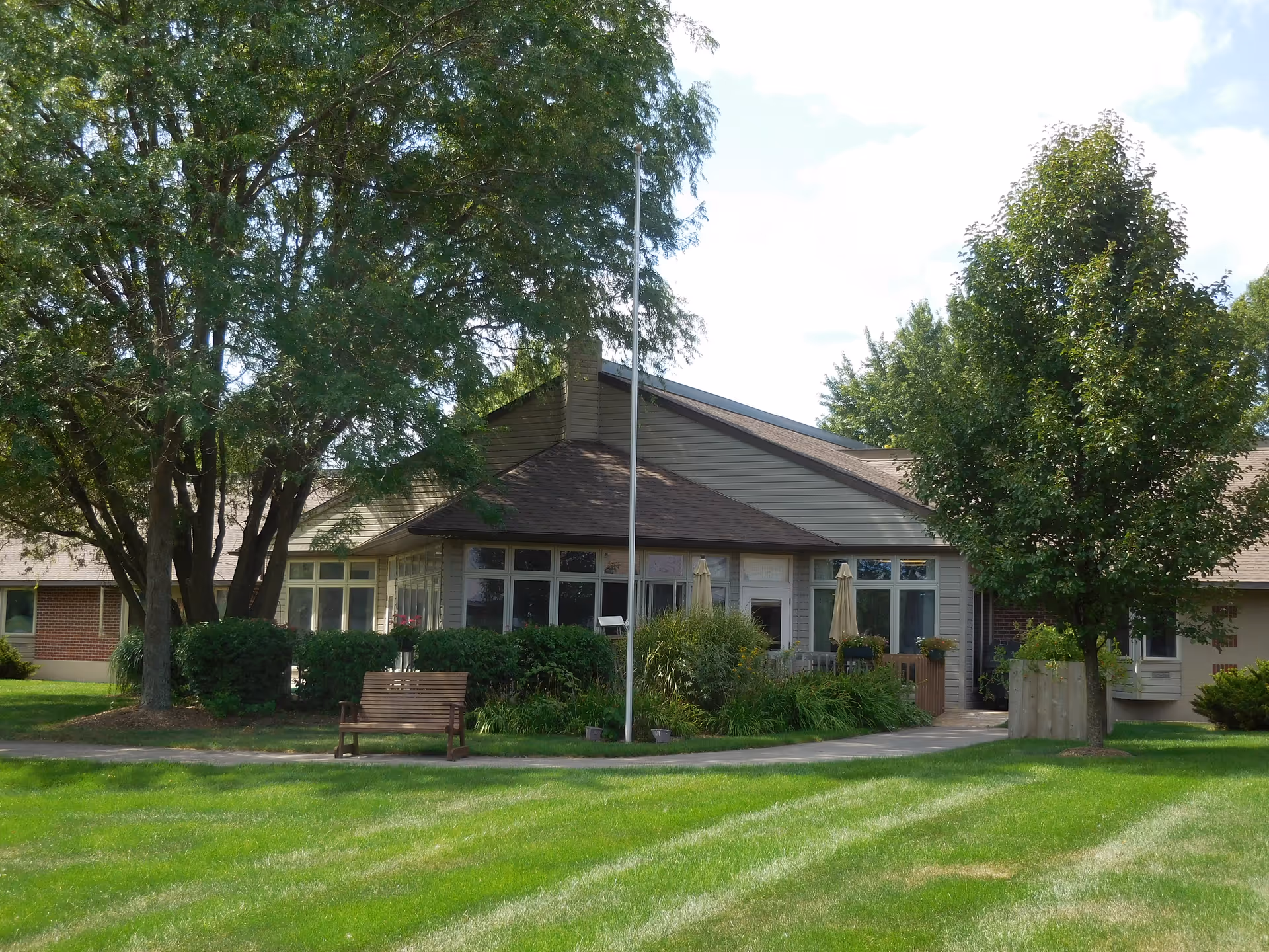 Single-story assisted living building front with large windows, trees, a bench, and a manicured lawn.