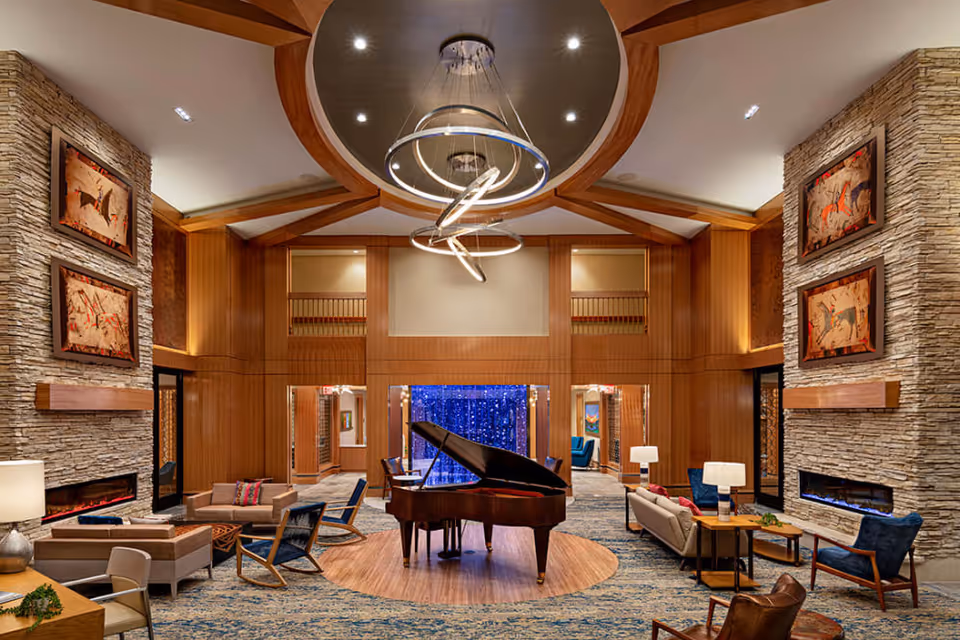 A spacious and elegant living room area in a senior living facility featuring a grand piano at the center on a circular wooden platform. The room has high ceilings with modern circular light fixtures, stone accent walls with framed artwork, multiple seating arrangements including sofas, armchairs, and rocking chairs, and a cozy fireplace on each stone wall. The decor includes warm wood paneling and a blue patterned carpet.