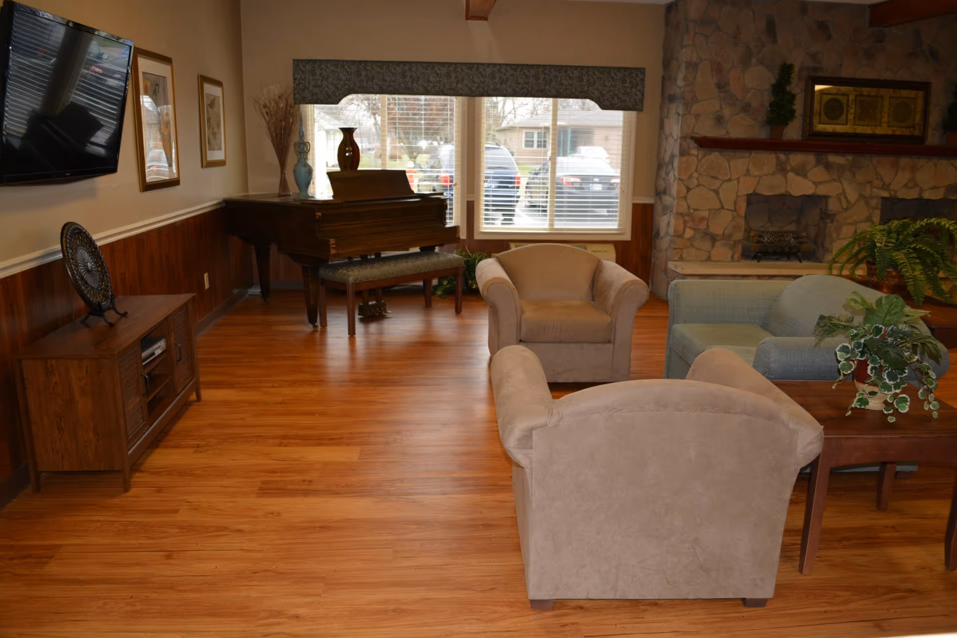 A cozy living room with wooden flooring, two beige armchairs, a light blue loveseat, a wooden coffee table with potted plants, a stone fireplace with framed artwork and small plants on the mantel, a wooden piano with decorative vases, a wall-mounted flat-screen TV, and large windows with blinds showing parked cars outside.
