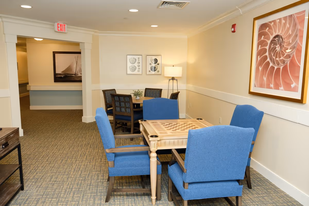 A cozy seating area in a senior living facility with a wooden game table featuring a checkerboard pattern surrounded by four blue upholstered chairs. In the background, there is another round table with four dark wooden chairs, a floor lamp, and framed artwork on the walls. The space has beige walls, carpeted flooring, and an exit sign above a doorway leading to a hallway.