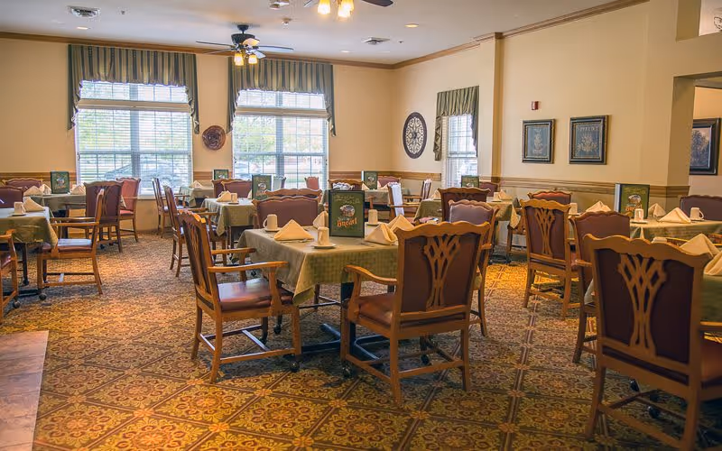 A dining room in a senior living facility with multiple tables covered with green tablecloths, each set with folded napkins and menus. Wooden chairs with cushioned seats surround the tables. The room has large windows with striped valances, framed artwork on the walls, and a patterned carpet.