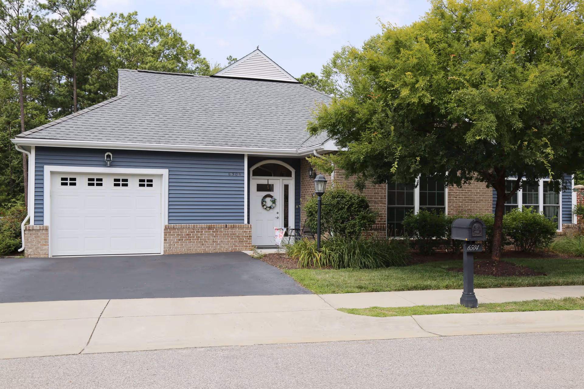 Exterior view of a single-story residential building with a gray shingled roof, blue siding, and brick accents. The building has a white garage door, a white front door with a wreath, a black mailbox on a post, a street lamp, and a large tree with green foliage in the front yard.