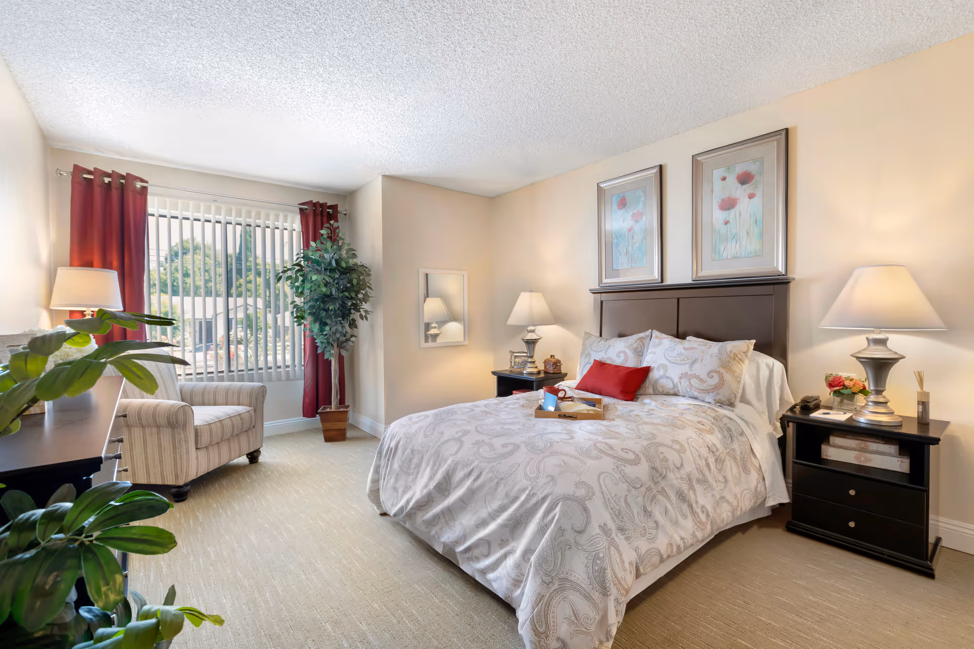 A bright and cozy bedroom with a large bed covered in a patterned white and gray comforter, two framed floral artworks above the headboard, two bedside tables with lamps, a striped armchair near a window with red curtains and vertical blinds, and several green plants adding a touch of nature to the room.