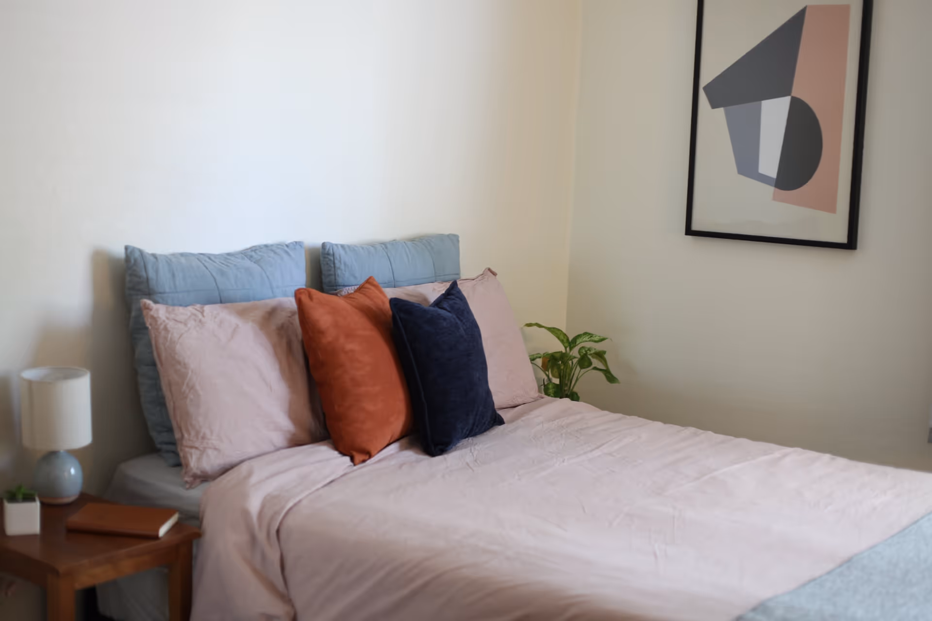 A neatly made bed with light pink sheets and multiple pillows in blue, pink, orange, and dark blue colors. There is a wooden nightstand with a small lamp, a small plant, and a closed book. A framed abstract artwork hangs on the wall above the bed, and a green plant is placed in the corner of the room.
