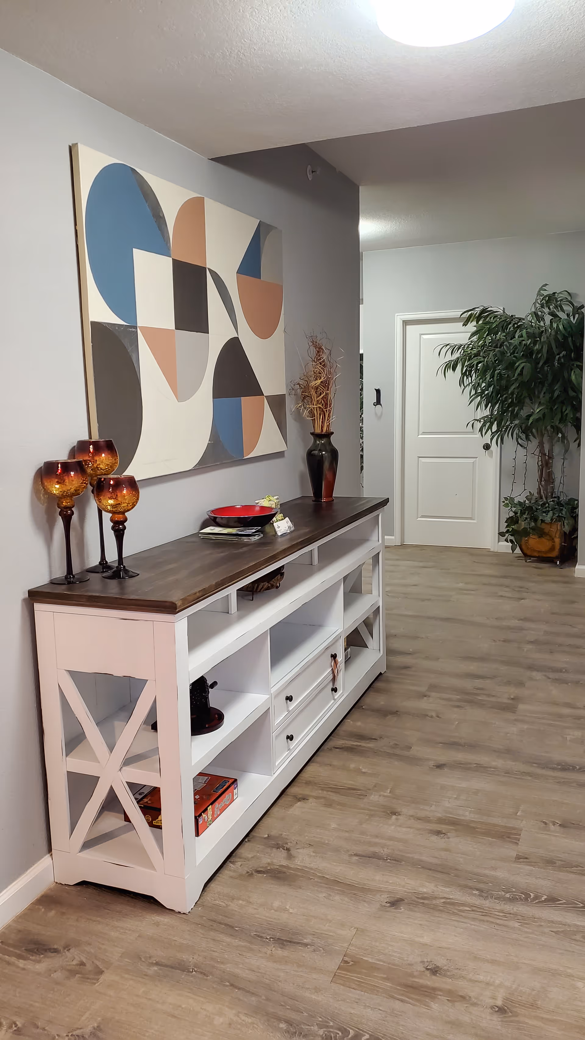 A hallway with a white console table featuring a dark wooden top. On the table are three amber-colored decorative glasses, a red bowl, and a tall black vase with dried branches. Above the table hangs a large abstract painting with geometric shapes in blue, beige, black, and brown. The floor is light wood, and a green potted plant is visible near a white door at the end of the hallway.