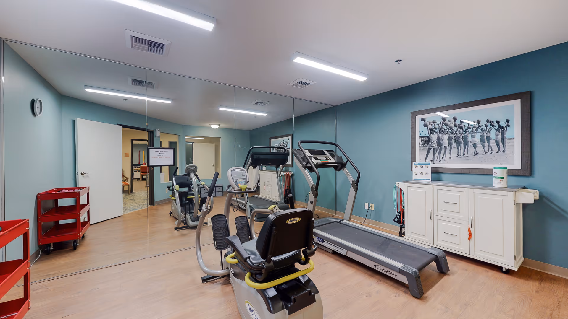 A small fitness room with light blue walls and wooden flooring. The room contains exercise equipment including a recumbent bike and a treadmill. One wall is covered with a large mirror reflecting the equipment and part of the room. There is a white cabinet against the wall with a framed black and white photo of people exercising above it. A red utility cart is positioned near the door, which is open to an adjacent room.