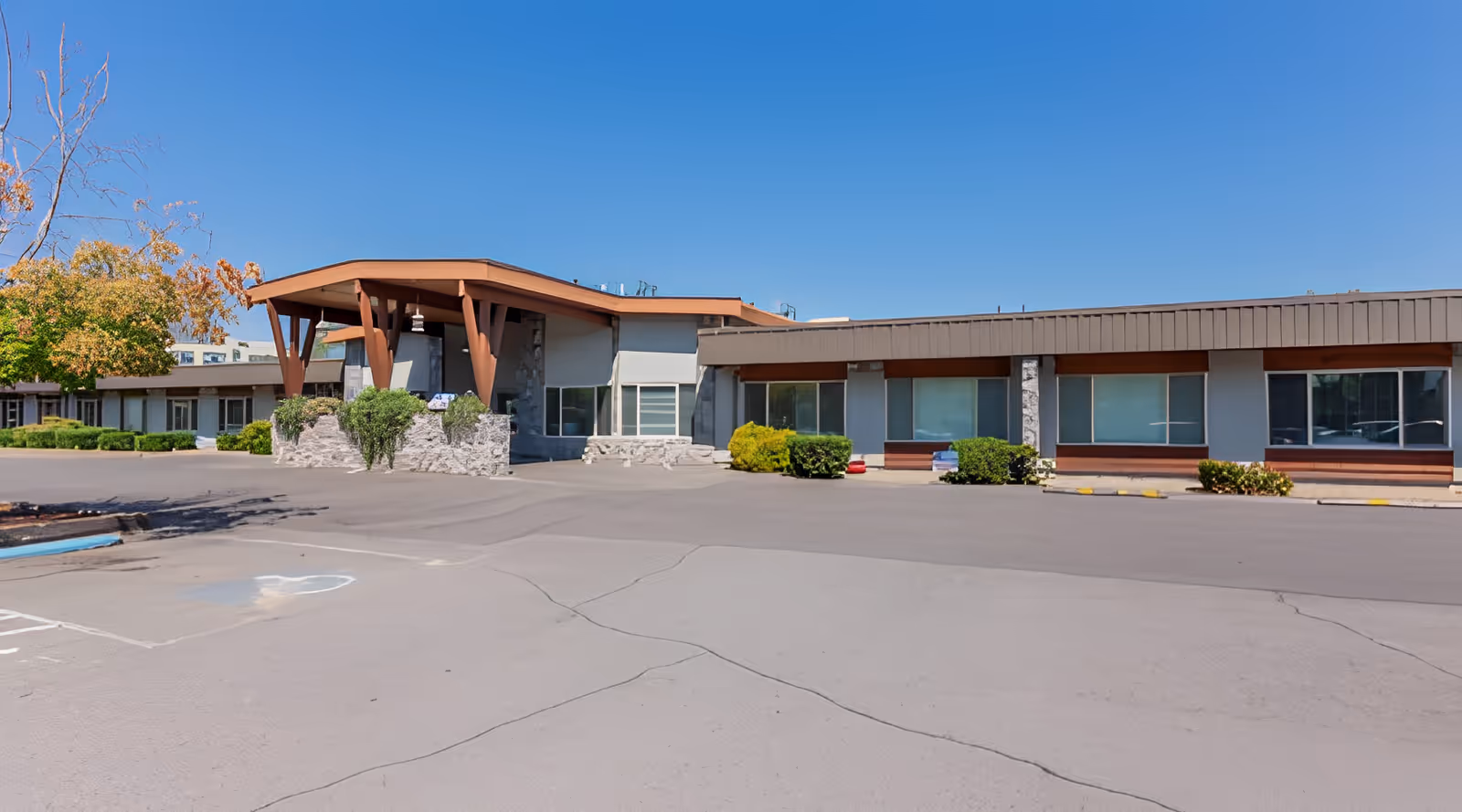 Exterior view of Hearthstone Nursing & Rehabilitation Center showing a single-story building with large windows, a covered entrance supported by wooden beams, surrounded by bushes and trees under a clear blue sky.