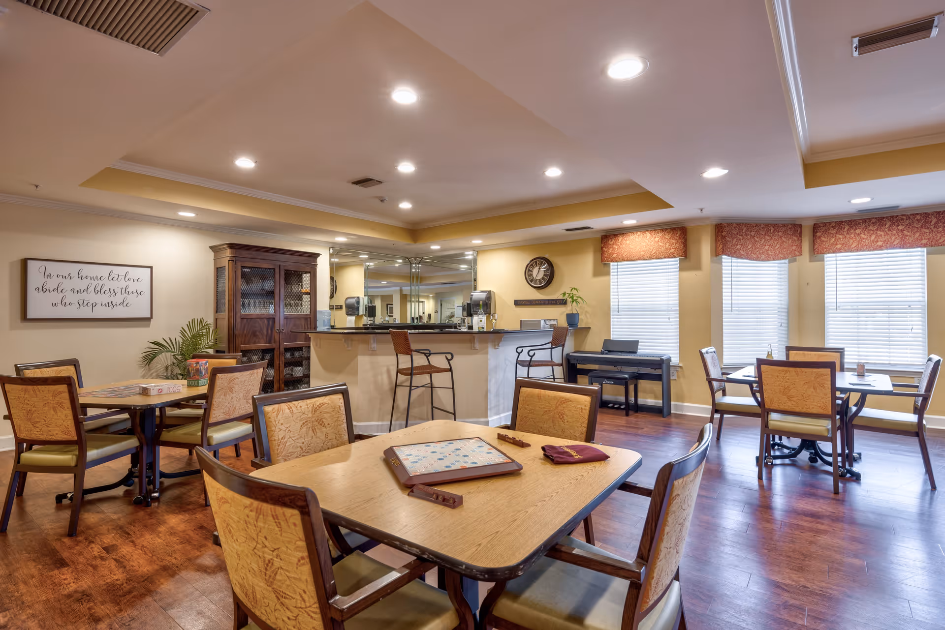 Bright common dining/activity room with multiple tables and chairs, a small service counter, and a piano by the windows.