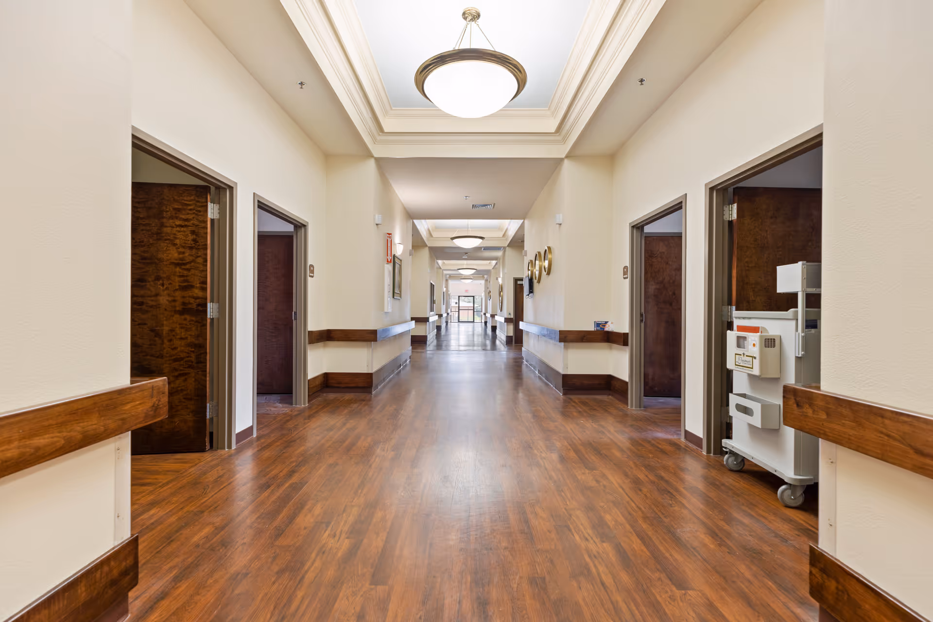 A long, well-lit hallway in a senior living facility with wooden floors and beige walls. The hallway has several open doorways on both sides, wooden handrails along the walls, and ceiling lights with decorative fixtures. A medical cart is visible near one of the doorways on the right side.