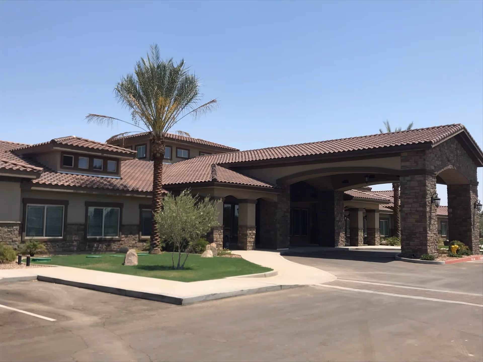Front exterior of a single-story memory care building with a covered porte-cochère, stone columns, tiled roof, and palm trees.