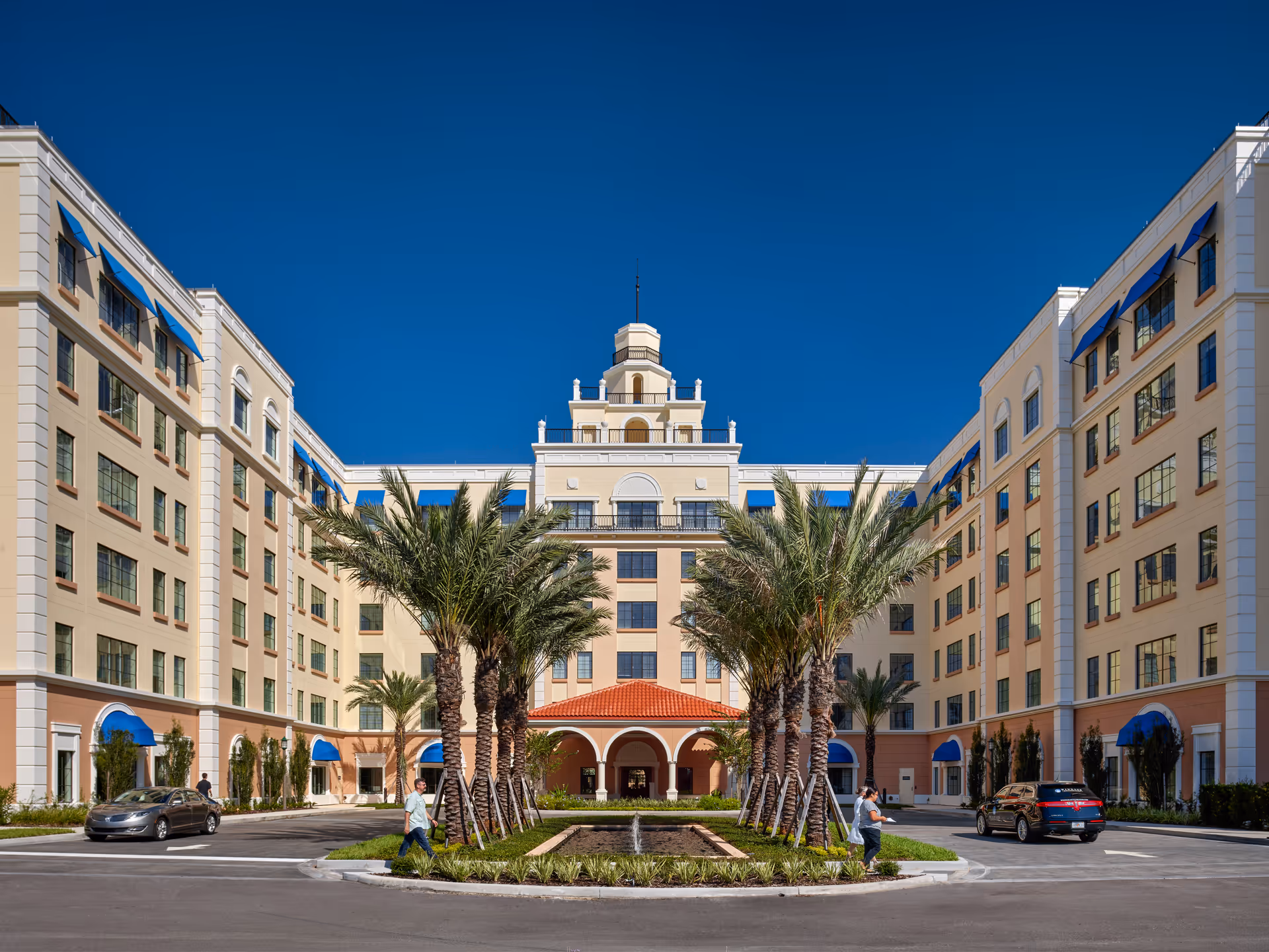 Front exterior of a multi-story senior living building with a central arched entrance, palm trees, and parked cars under a clear blue sky.