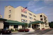 Exterior view of a multi-story beige building with a green roof entrance and an American flag flying on a flagpole in front. Several cars are parked near the entrance under a clear sky.