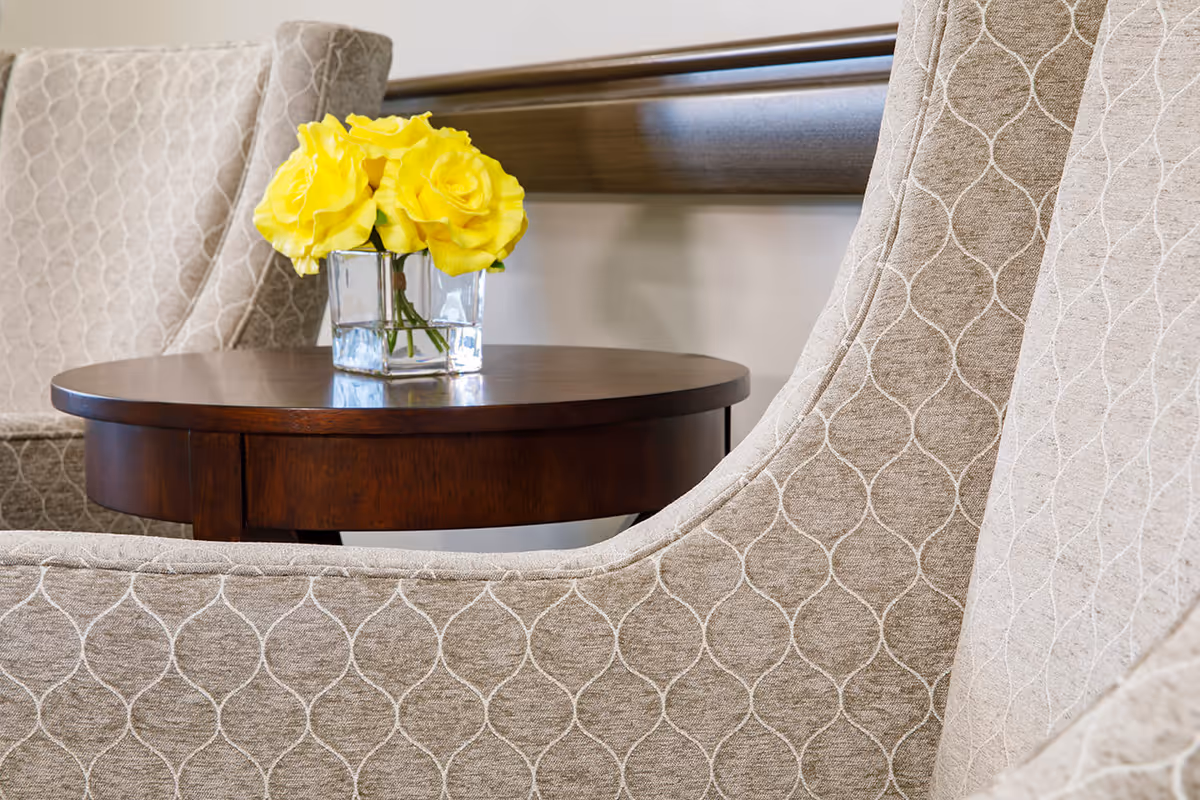 Close-up view of a small round wooden table with a clear glass vase holding bright yellow roses, positioned between two beige upholstered chairs with a subtle geometric pattern.