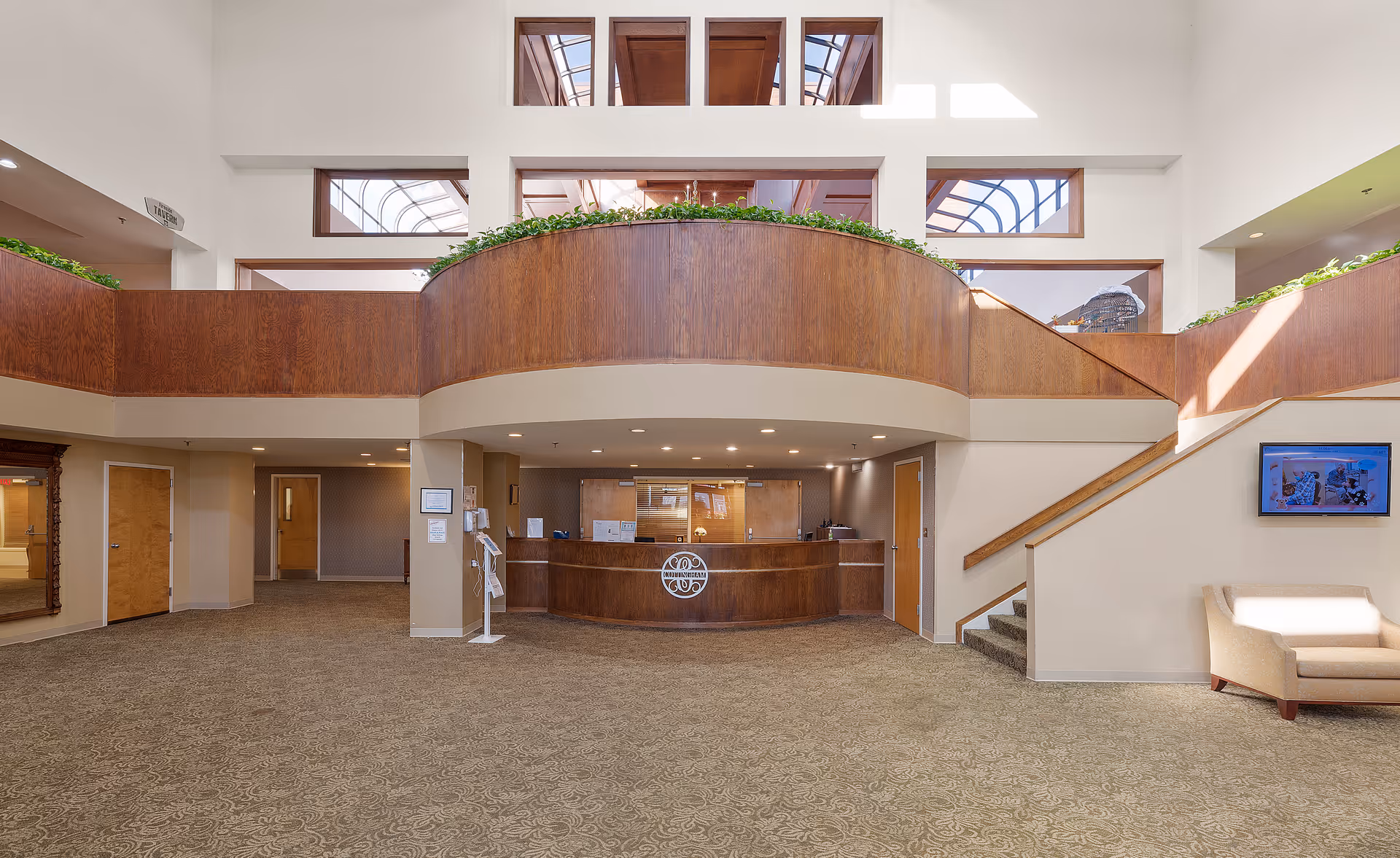 Spacious and well-lit reception area of Cottingham Retirement Community featuring a wooden front desk with the community logo, carpeted floor with intricate patterns, a staircase on the right, large windows above allowing natural light, and a beige armchair near a wall-mounted television.