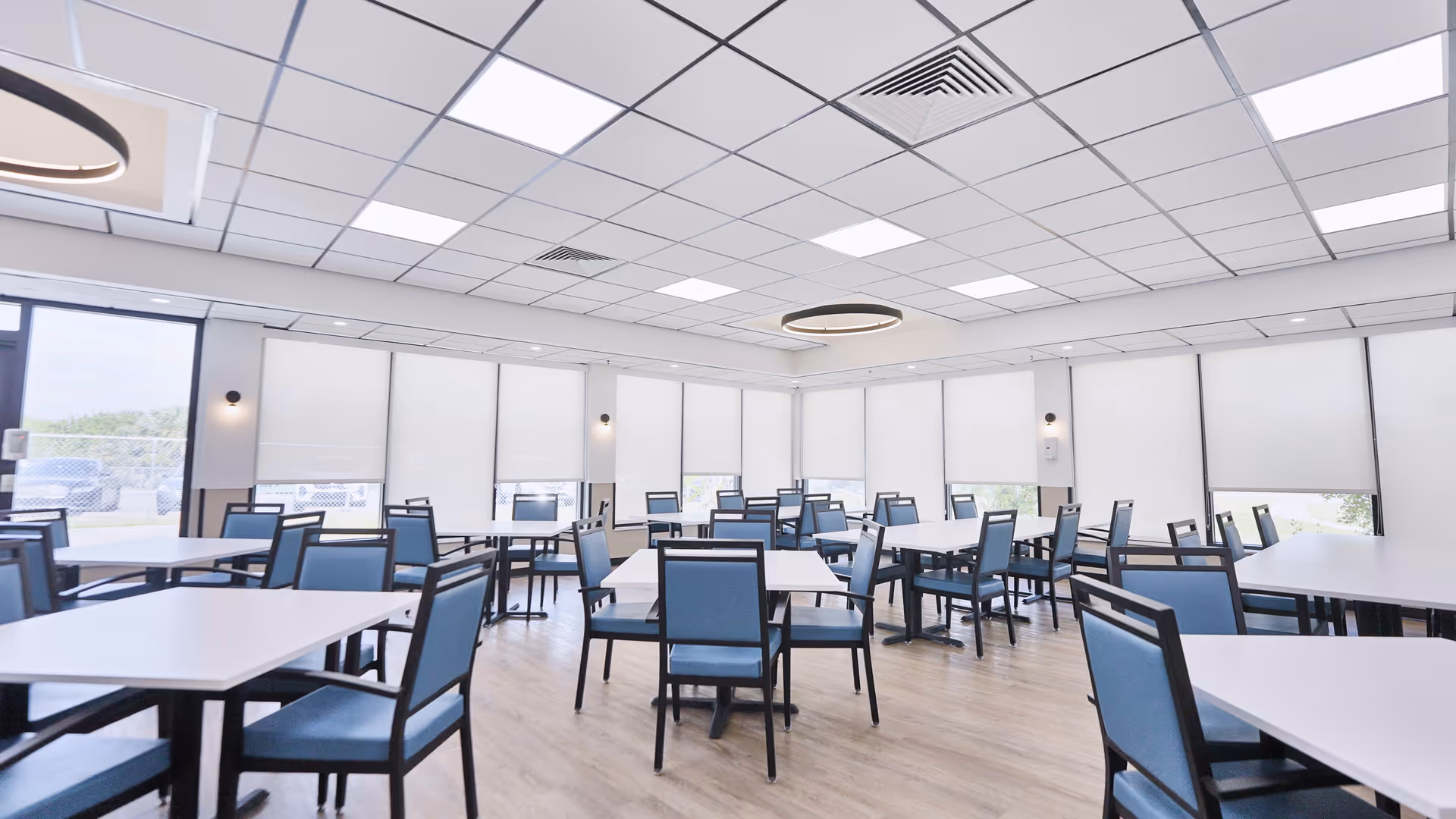 A bright and spacious dining room with multiple white tables and blue cushioned chairs arranged neatly. Large windows with white roller blinds cover the walls, allowing natural light to filter in. The ceiling has a grid pattern with recessed lighting and a modern circular light fixture in the center. The floor is wooden, and the room appears clean and well-organized.
