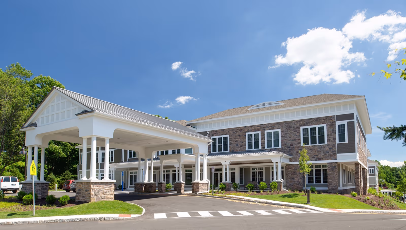 Exterior view of a large, modern senior living facility building with stone and siding facade, multiple windows, and a covered drop-off area supported by white columns. The sky is clear with a few clouds, and there is landscaping with grass and small bushes around the building.