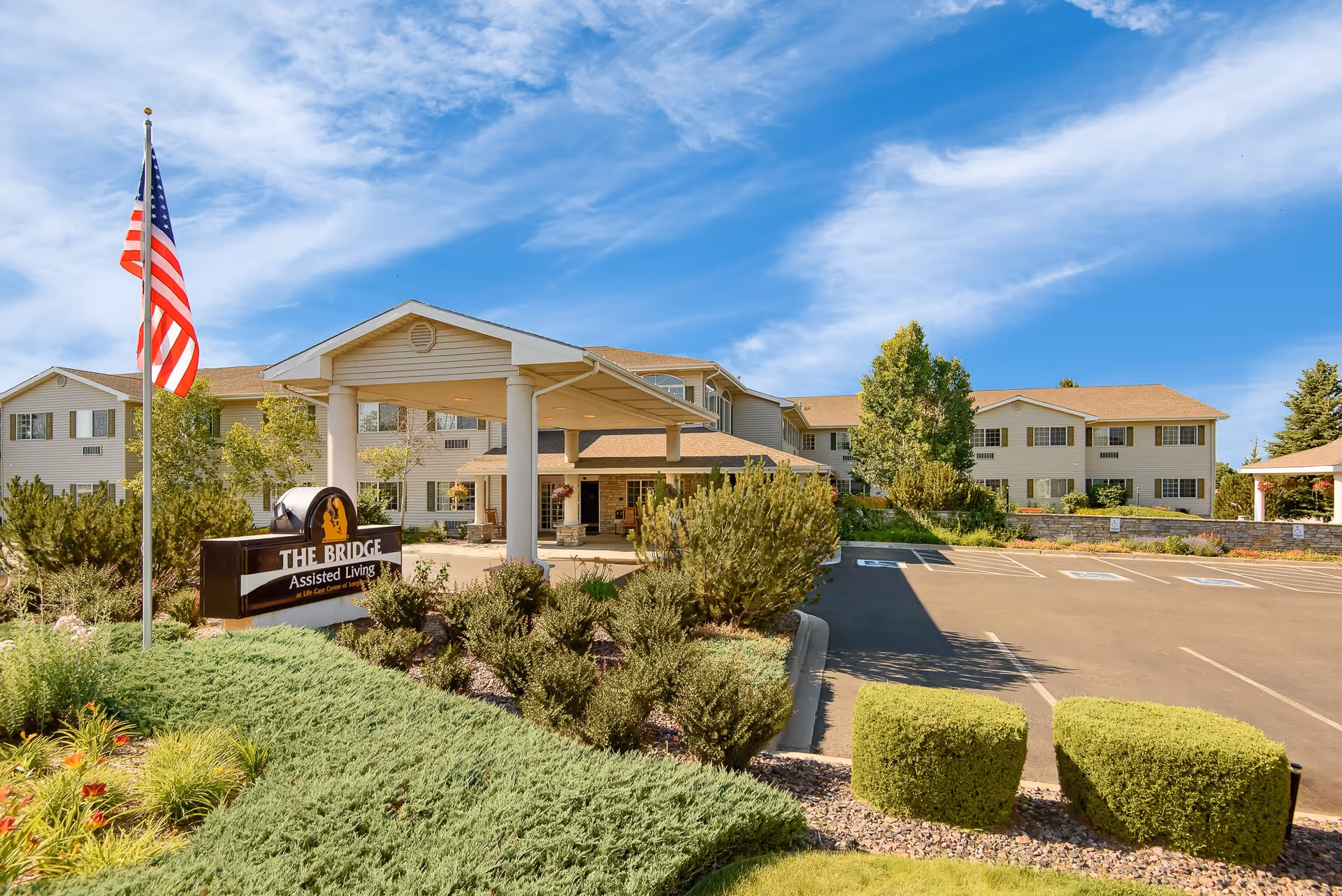 Exterior view of The Bridge at Longmont assisted living facility showing a large two-story building with beige siding and a covered entrance. There is a landscaped garden with bushes and flowers in the foreground, an American flag on a flagpole, and a parking lot with several empty spaces under a blue sky with scattered clouds.